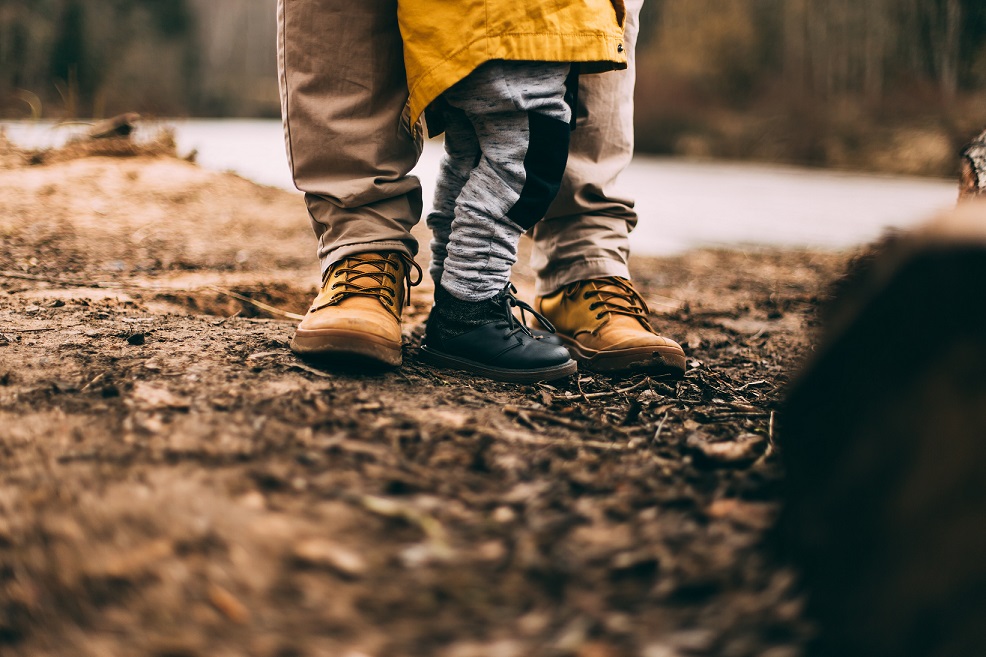 The legs and feet of a child and adult are visible standing close together in a natural setting