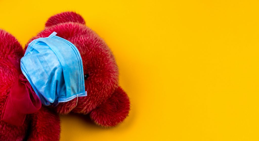 A red teddy bear against a yellow background wearing a blue surgical mask