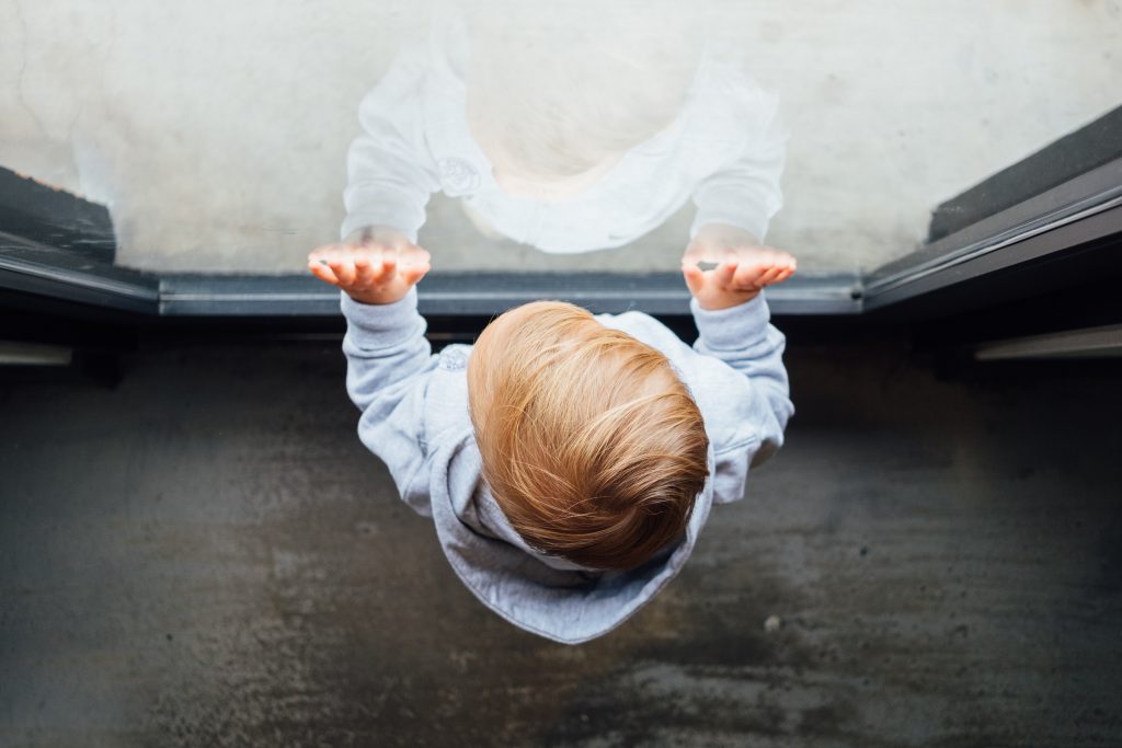 A young child looks out a window with their hands placed on the windowpane