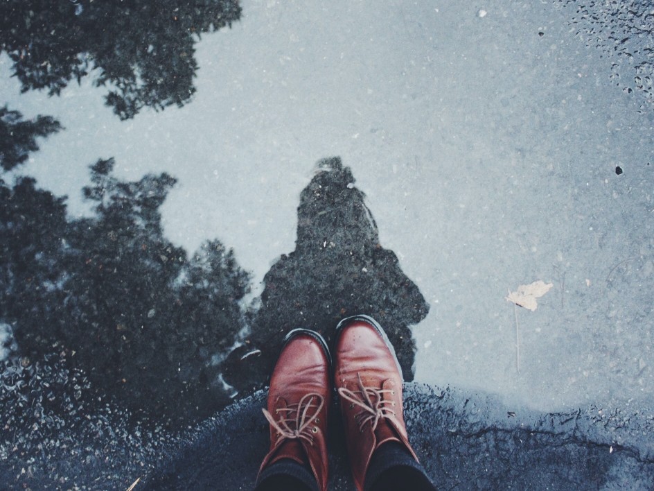 A puddle reflecting the sky, tree branches and a person whose boots are seen at the bottom of the photo