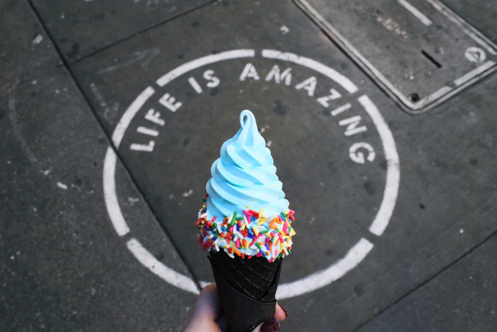 A person's hand holds an ice cream cone with sprinkles in front of the words "Life is amazing" painted on the street