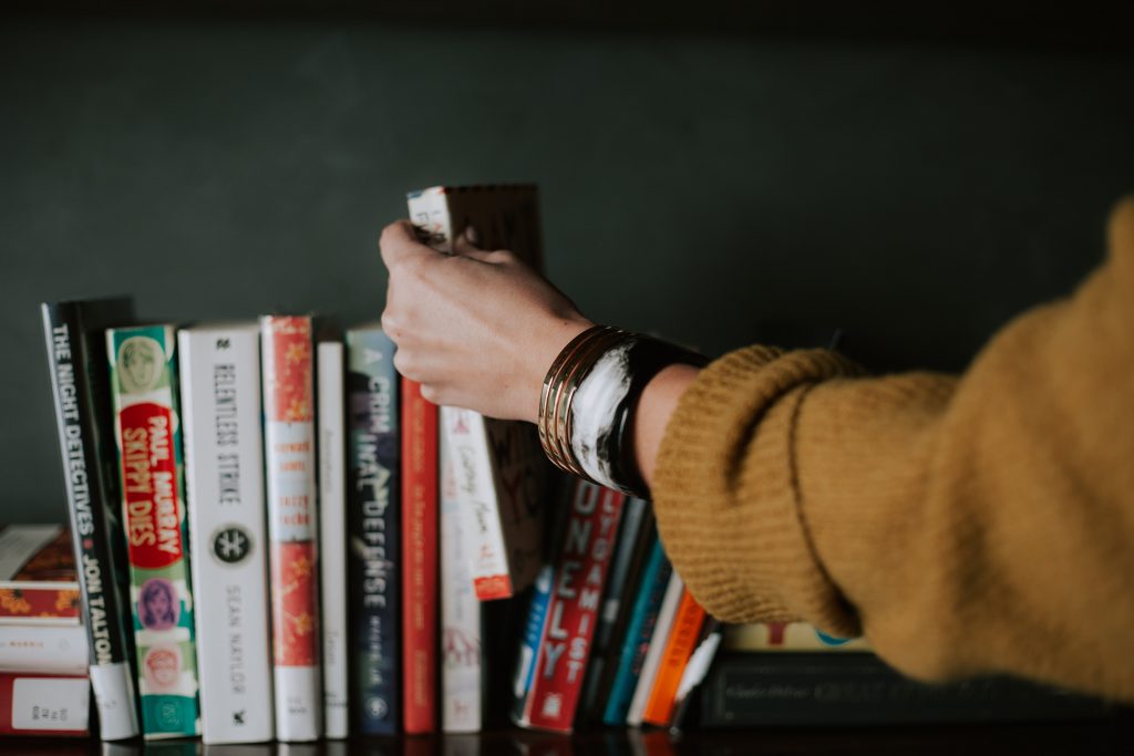 A person wearing bracelets and a yellow sweater chooses a book from a row of books on a shelf