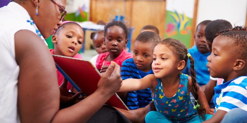 A group of children look intently at a book held by an adult. One child leans forward to point at the page