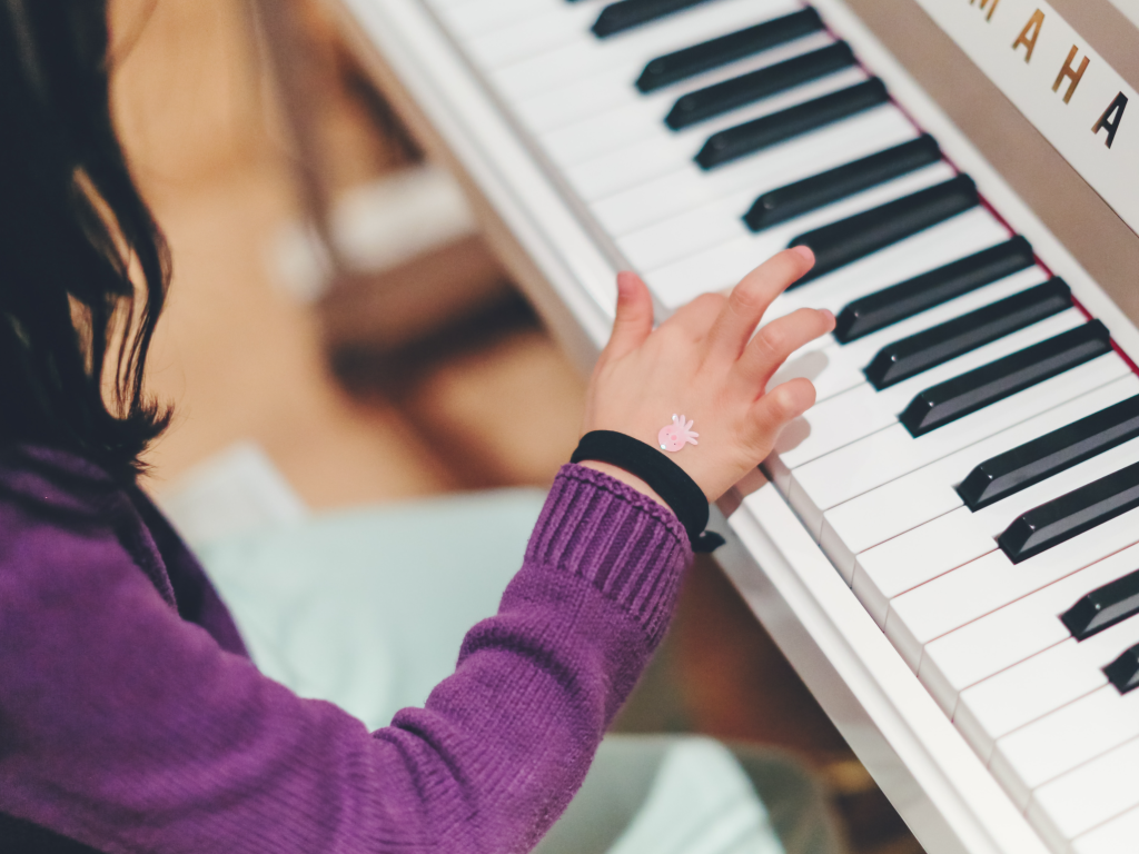 A child wearing a purple sweater with a small pink sticker on the back of her hand presses a key on a piano keyboard with her index finger.