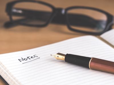 A pen on a notebook where the word "notes" is written and a pair of glasses in the background
