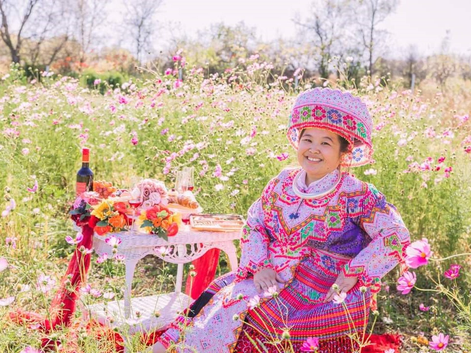 Barbara Vang in traditional Hmong dress at a table covered with flowers, food and drinks in a sunny field of wildflowers