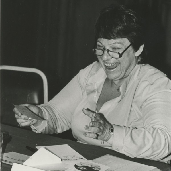 Erna Fishhaut sits at a table covered with papers, her arms outstretched, wearing glasses, a white shirt, and a big smile