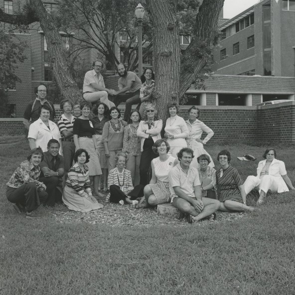 Attendees of the 1977 Professional Growth Institute sit or stand on the grass and in or around a large tree outside university buildings