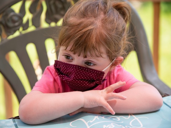 A young girl with red hair and light-toned skin sits with arms folded on a tabletop wearing a polka-dot cloth face mask